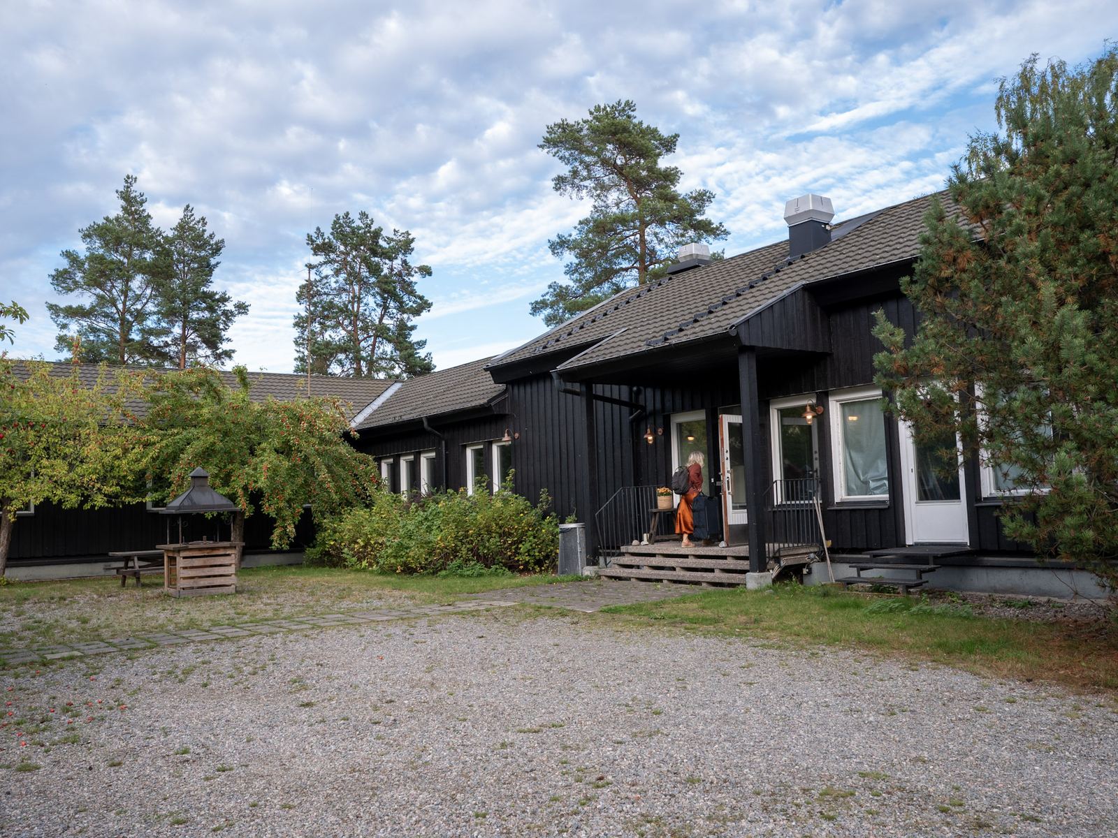 Gårdens mörka trähus med entrén, bland höga tallar.